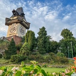 Grazer Uhrturm erhebt sich über üppigen Gärten mit bunten Blumen. Der historische Turm zeigt zwei große Zifferblätter unter blauem Himmel.