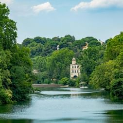 Canal du Midi near Béziers with a historic tower surrounded by lush green trees. A boat is visible on the calm water under a blue sky.