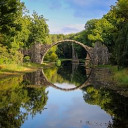 Historische Steinbogenbrücke Rakotzbrücke in Kromlau spiegelt sich perfekt im ruhigen Wasser und bildet einen Kreis mit grünem Wald.