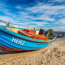 Buntes Fischerboot mit blauem Rumpf HER2 am sandigen Ostseestrand. Orange Netze und Ausrüstung im Inneren unter blauem Himmel sichtbar.