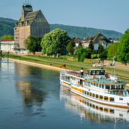 White paddle steamer with German flag on Weser River in Holzminden. Historic grain silo and residential buildings visible on riverbank with hills behind.