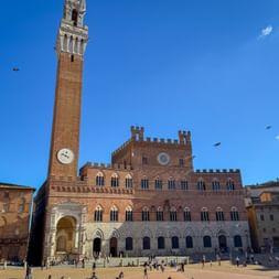 Piazza del Campo in Siena with the medieval Palazzo Pubblico and its tall Torre del Mangia tower against a blue sky. Visitors gather on the square.