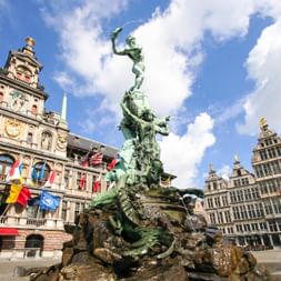 Brabo-Brunnen aus Bronze auf dem Grote Markt in Antwerpen, mit historischen Gildehäusern und dem verzierten Rathaus mit Flaggen im Hintergrund.