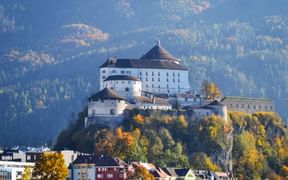 Kufstein Fortress perched on a hill above colorful buildings in autumn. The medieval fortress overlooks the town with forested mountains in background.