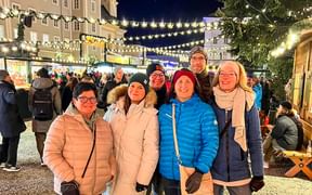 Six team members in winter clothing smiling at a Christmas market with festive lights and market stalls in the evening.