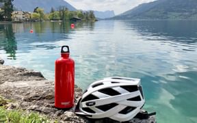 Red water bottle and white cycling helmet on rocky shore of alpine lake with mountains and village in background, Salzkammergut region.