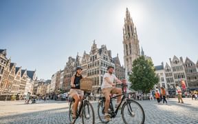 Two cyclists riding on cobblestone Grote Markt square in Antwerp, with historic guild houses and cathedral tower in background under sunny sky.