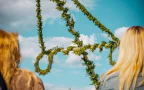 Zwei Personen mit blonden Haaren betrachten einen geschmückten Mittsommerbaum von unten vor blauem Himmel mit weißen Wolken.