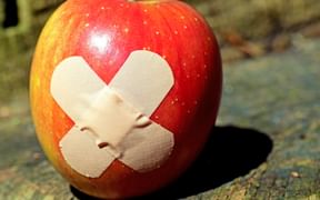 Red and yellow apple with two crossed bandages on its surface, placed on weathered wooden surface in sunlight.