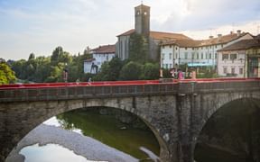 Stone arch bridge with red railings crossing a river in Cividale del Friuli. Historic buildings and a church tower visible in the background.
