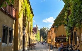 Cobblestone street in Augsburg's Fuggerei with yellow row houses covered in ivy, outdoor seating, and cyclists under blue sky.