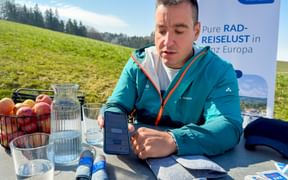 Man in turquoise jacket at outdoor table with phone, water, and fruit basket. Eurobike banner visible behind him in green alpine landscape.