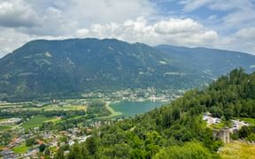 Panoramablick von Landskron über den Ossiachersee in Kärnten mit bewaldeten Hügeln, einem Dorf und Bergen unter bewölktem Himmel.