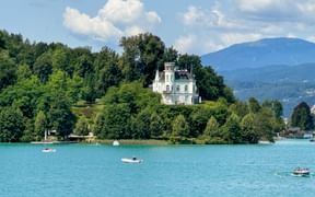 Weißes Schloss Reifnitz auf grünem Hügel mit Blick auf den türkisfarbenen Wörthersee mit Booten, Bergen im Hintergrund unter blauem Himmel.