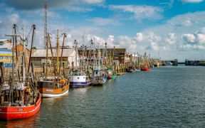 Cuxhaven harbor with colorful fishing boats moored along the quay. Red, orange and blue vessels line the waterfront with industrial buildings.