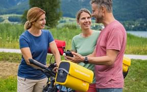 Three cyclists with e-bikes checking a route app by Wolfgangsee lake in Salzkammergut. Mountains and green meadows visible in background.