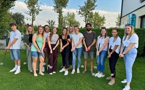 Group of 13 young people standing on grass holding golf clubs, smiling at camera. Trees and white building visible in background.