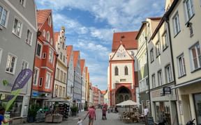 Cobblestone pedestrian street in Landsberg with colorful historic buildings and a medieval tower gate. People walk and dine at outdoor cafes.