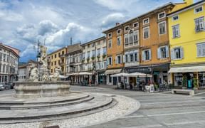 Neptune fountain in Gorizia's old town square surrounded by colorful historic buildings. Cafes line the piazza under a dramatic cloudy sky.