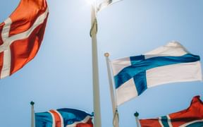 Mehrere nordische Flaggen, darunter dänische, schwedische, finnische und isländische Flaggen, wehen vor einem klaren blauen Himmel mit hellem Sonnenlicht.