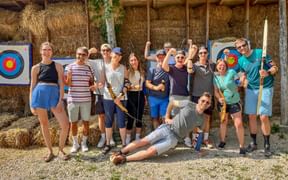 Group of people posing with bows and arrows at outdoor archery range with hay bale targets and wooden shelter structure.