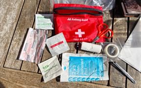 Red first aid kit with white cross and cycling tour supplies including bandages, scissors, tape, and antiseptic wipes on wooden surface.
