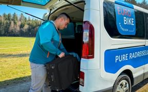 Mann in türkisfarbener Jacke lädt schwarzen Koffer in weißen Eurobike-Transporter mit offenen Hecktüren in sonniger Landschaft.