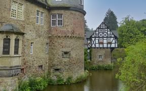 Historic Crottorf water castle with stone tower and half-timbered building reflected in surrounding moat, surrounded by lush green trees.