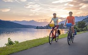 Zwei Radfahrer fahren entlang eines Uferwegs am Millstätter See in Kärnten bei Sonnenuntergang, mit Bergen im Hintergrund und ruhigem Wasser.