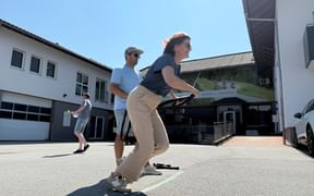 Woman in beige pants and dark shirt playing an outdoor game in a courtyard, with a man watching behind her and white buildings visible.