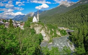 Scenic view of Scuol village with white church on rocky hill, Inn River flowing through valley, surrounded by forested mountains under blue sky.