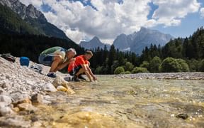 Two hikers crouching by the clear Pisnica River in Slovenia, touching the water. Mountain peaks and forests visible in the background.