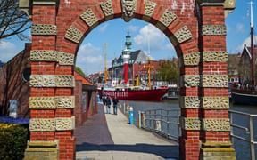 Red brick archway with decorative stonework frames view of Emden harbor. Red lightship and historic buildings visible through arch under blue sky.