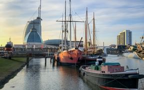 Historic sailing ships moored in Bremerhaven museum harbor with modern buildings and glass dome in background under cloudy sky.