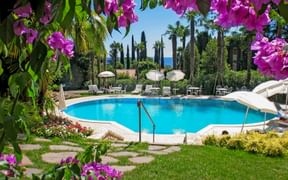 Blauer Swimmingpool der Villa Sofia in Gardone, eingerahmt von rosa Bougainvillea. Weiße Sonnenliegen und Schirme umgeben den Pool mit Palmen und Seeblick.