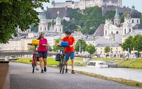Zwei Radfahrer gehen mit Rädern an der Salzach in Salzburg entlang. Festung Hohensalzburg und historische Kirchen im Hintergrund.