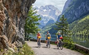 Three cyclists on lakeside path at Gosausee with Dachstein mountain range in background. Rocky cliff on left, alpine lake and forested slopes visible.