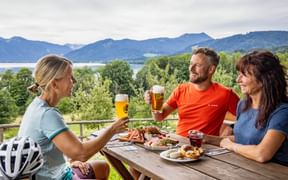 Three cyclists toasting with beer at a wooden table on a terrace. Lake Tegernsee and mountains visible in the background. Food platters on the table.