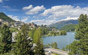 Panoramic view of St. Moritz with lake, snow-capped Alpine mountains, historic buildings on hillside, and blue sky with white clouds.