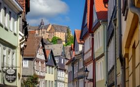 Enge Kopfsteinpflasterstraße in Marburgs Altstadt mit bunten Fachwerkhäusern und dem Landgrafenschloss auf dem Hügel.