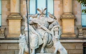 Steinstatue von Odin auf einem Thron sitzend, einen Speer haltend, flankiert von zwei Wölfen am Sockel und zwei Raben auf seinen Schultern, vor einem Sandsteingebäude.