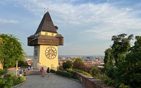 Der berühmte Grazer Uhrturm mit seinem charakteristischen Holzdach und großen Zifferblatt, mit Blick über die Stadt Graz und Besuchern auf der Terrasse.