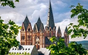 Historischer Limburger Dom mit charakteristischen sieben Türmen aus rotem Sandstein und dunklen Turmspitzen, umrahmt von grünem Laub.