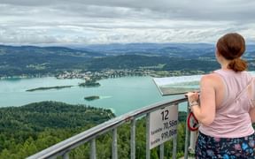 Frau auf der Aussichtsplattform Pyramidenkogel mit Blick auf den Wörthersee in Kärnten mit bewaldeten Hügeln und bewölktem Himmel.