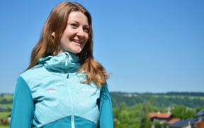 Smiling woman wearing a turquoise Eurofun outdoor jacket standing outdoors against a blue sky with green landscape in the background.