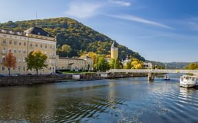 Bad Ems Flussufer mit historischem Rundturm, gelben Barockgebäuden, Brücke und Booten auf der Lahn mit bewaldeten Hügeln.