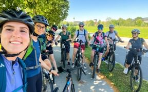 Group of cyclists with helmets posing on a paved path in Salzburg. Green fields and trees visible in background under blue sky.