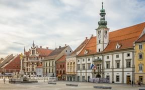Historischer Hauptplatz in Maribor mit bunten Barockgebäuden, einem markanten Kirchturm mit grüner Kuppel und einem zentralen Brunnen.
