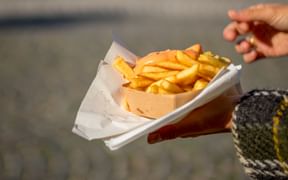 Hand hält eine Papiertüte mit goldenen belgischen Pommes auf einem Marktplatz in Flandern, Kopfsteinpflaster im Hintergrund sichtbar.