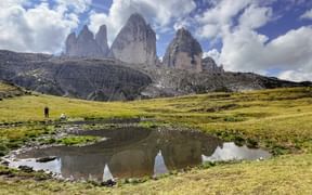 Die ikonischen Drei Zinnen Bergspitzen spiegeln sich in einem kleinen Teich, umgeben von grünen Almwiesen unter dramatischem Himmel.